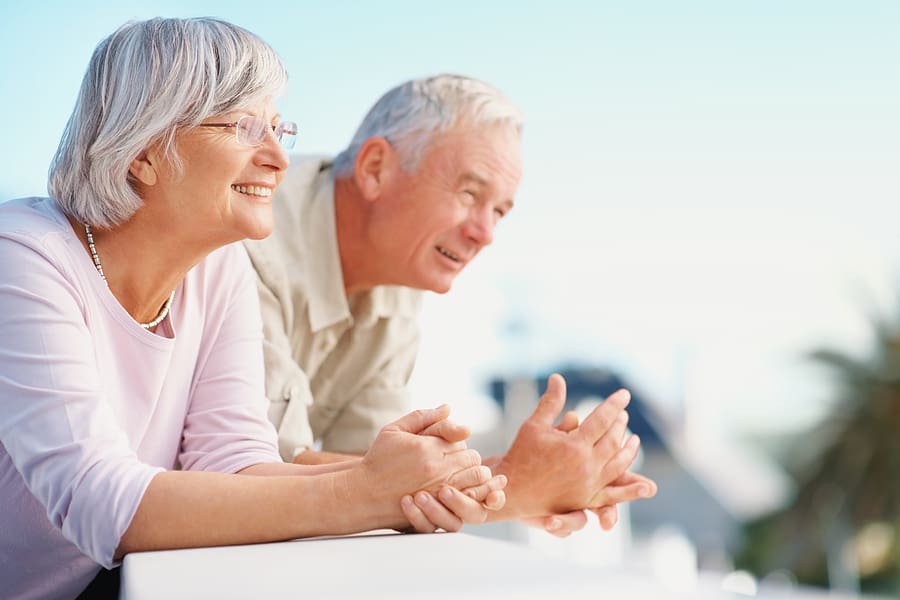 An elderly couple stands close together, smiling gently as they look ahead