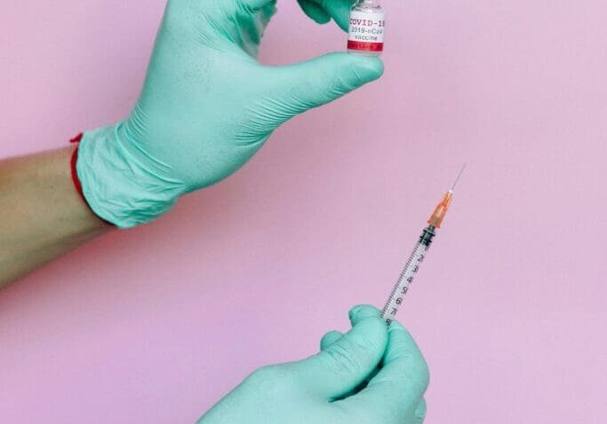 The image shows gloved hands, one is holding the vial and the other is holding an injection.