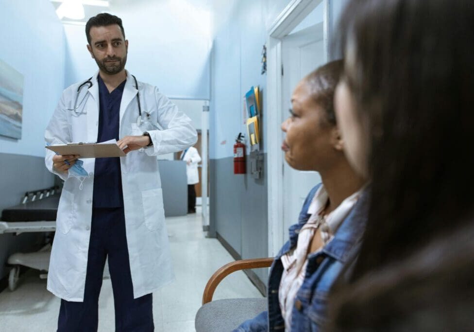 A Healthcare professional in a white coat discussing medical charts with a patient in a hospital hallway.