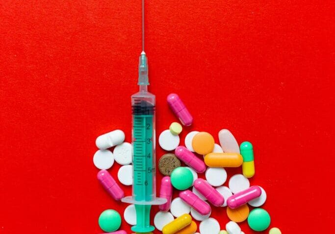 The image shows an injection surrounded by pills on a red background.