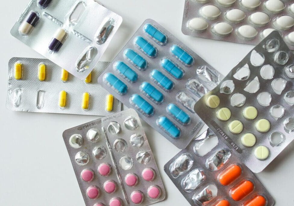 The image shows a variety of blister-packed medications, including blue, yellow, and pink capsules and tablets.